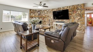 A living room with a stone wall, a ceiling fan, a flat-screen TV, and leather sofas. - Private Hot Tub & Huge Yard: Phoenix Family Home (Phoenix)