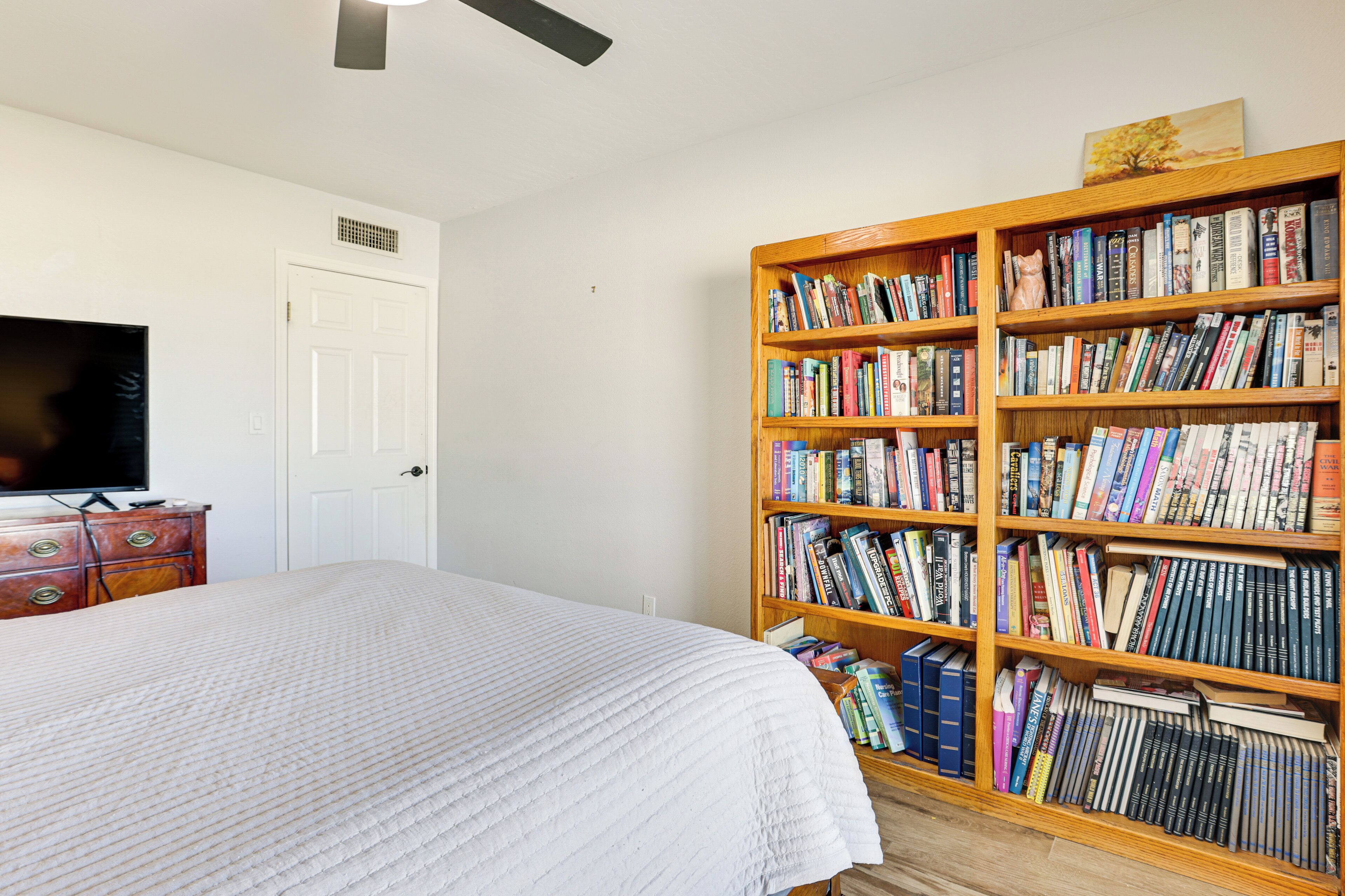 A bedroom with a large bed, a wooden bookshelf filled with books, a television on a dresser, and a white door.