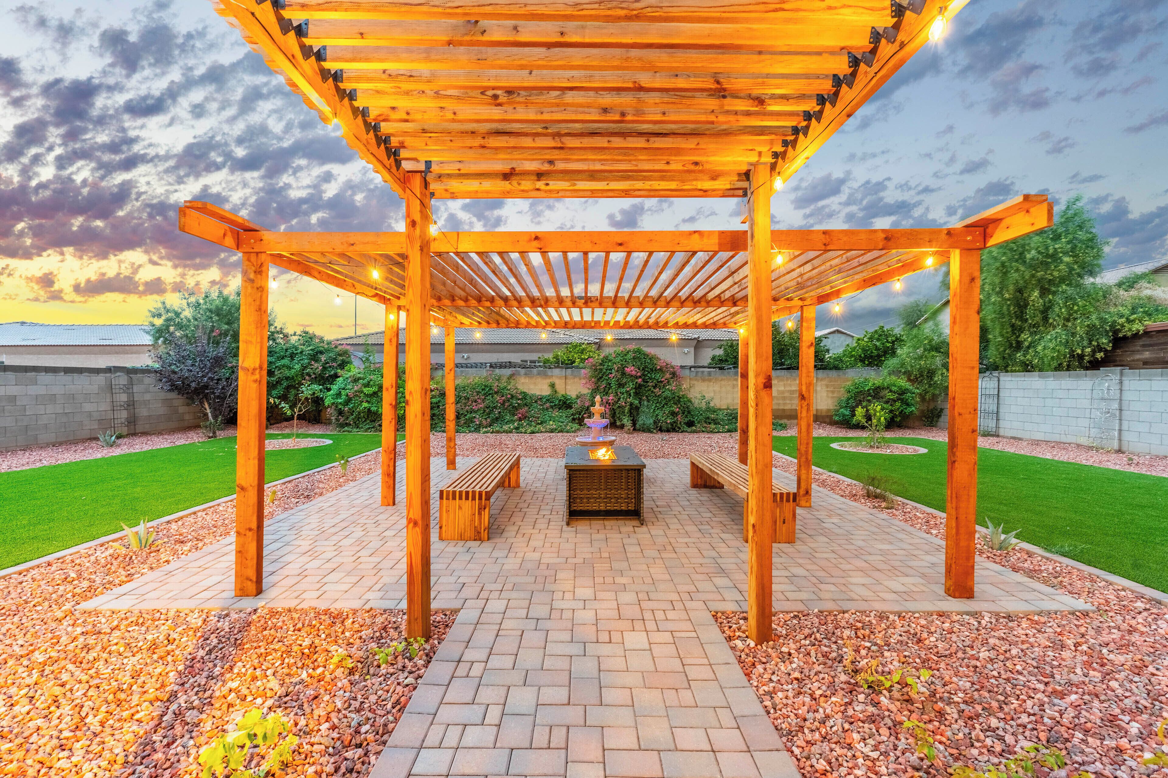 A wooden pergola with string lights, a fire pit, and benches in a landscaped garden area.