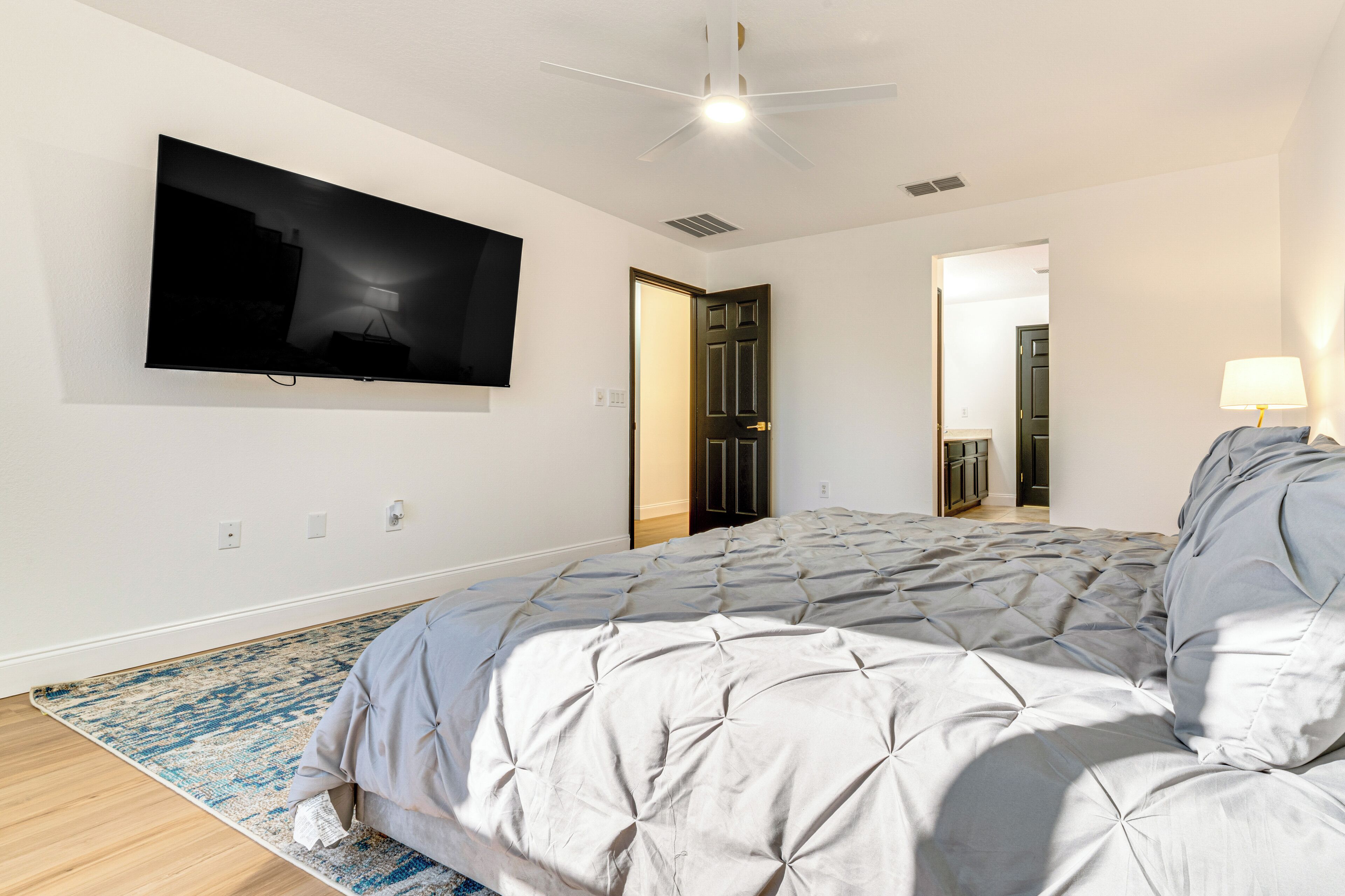 Un dormitorio con una cama, un televisor de pantalla plana en la pared, un ventilador de techo y una puerta que da a otra habitación.