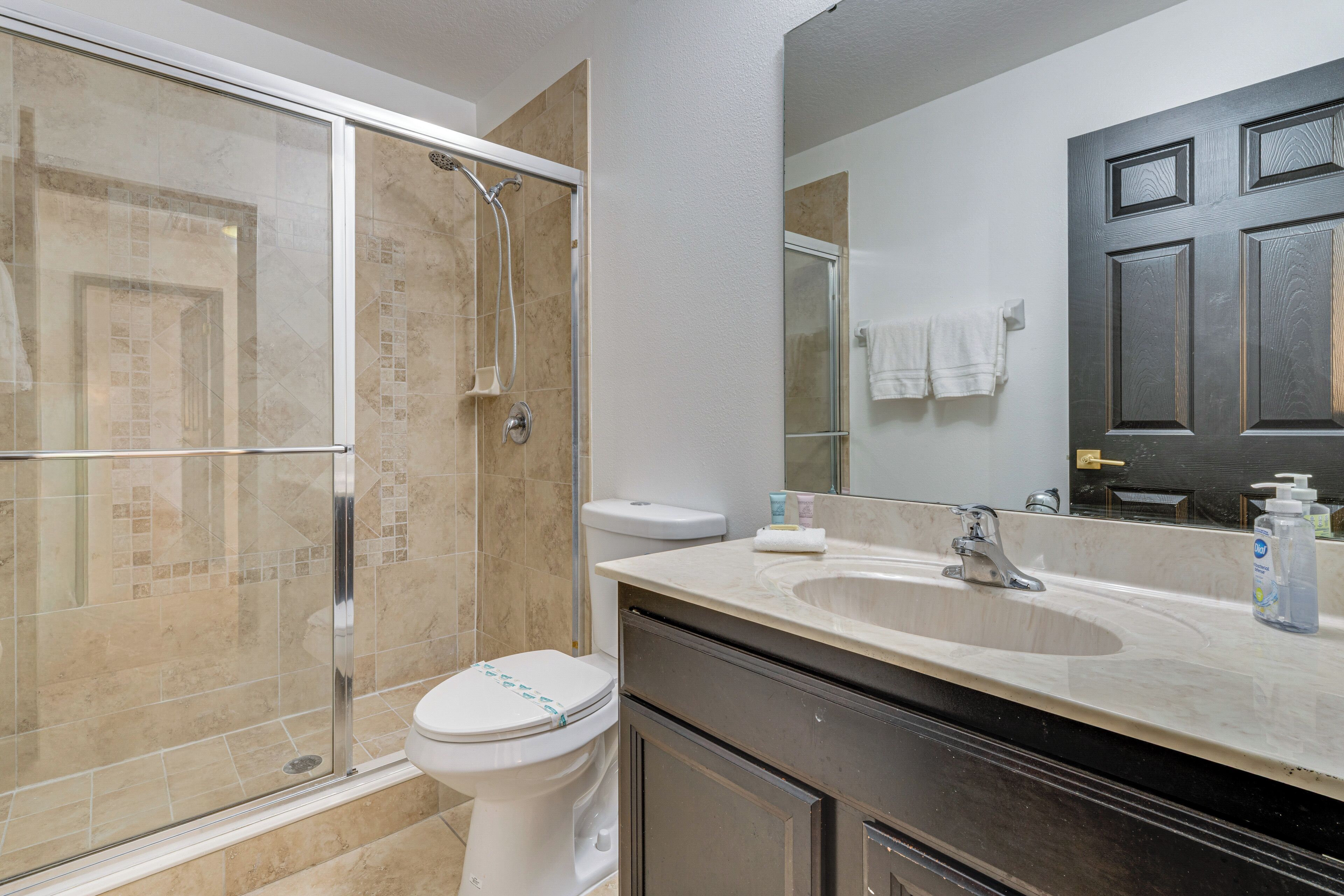 A bathroom with a glass-enclosed shower, a white toilet, a sink with a mirror, and a dark door.