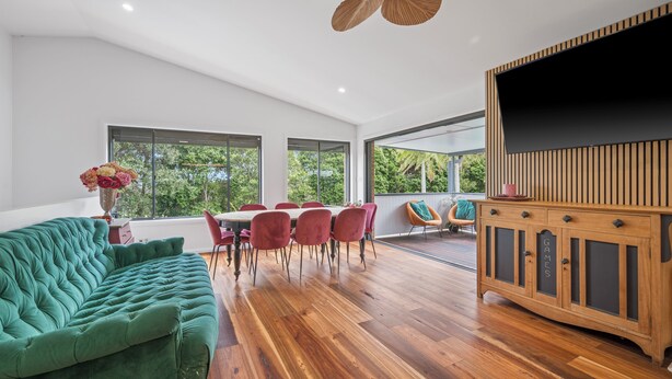 A spacious room with a green sofa, a dining table with red chairs, a wooden cabinet, and large windows offering a view of greenery. - Lighthouse Beach Coastal Escape (Port Macquarie)