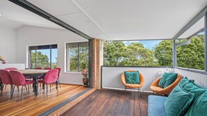 A modern interior with a wooden dining table and chairs, a blue sofa with teal cushions, and large windows offering a view of trees. - Lighthouse Beach Coastal Escape (Port Macquarie)