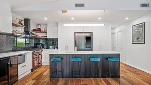 A modern kitchen with white cabinetry, a central island with bar stools, a built-in oven, and a stainless steel refrigerator. - Lighthouse Beach Coastal Escape (Port Macquarie)