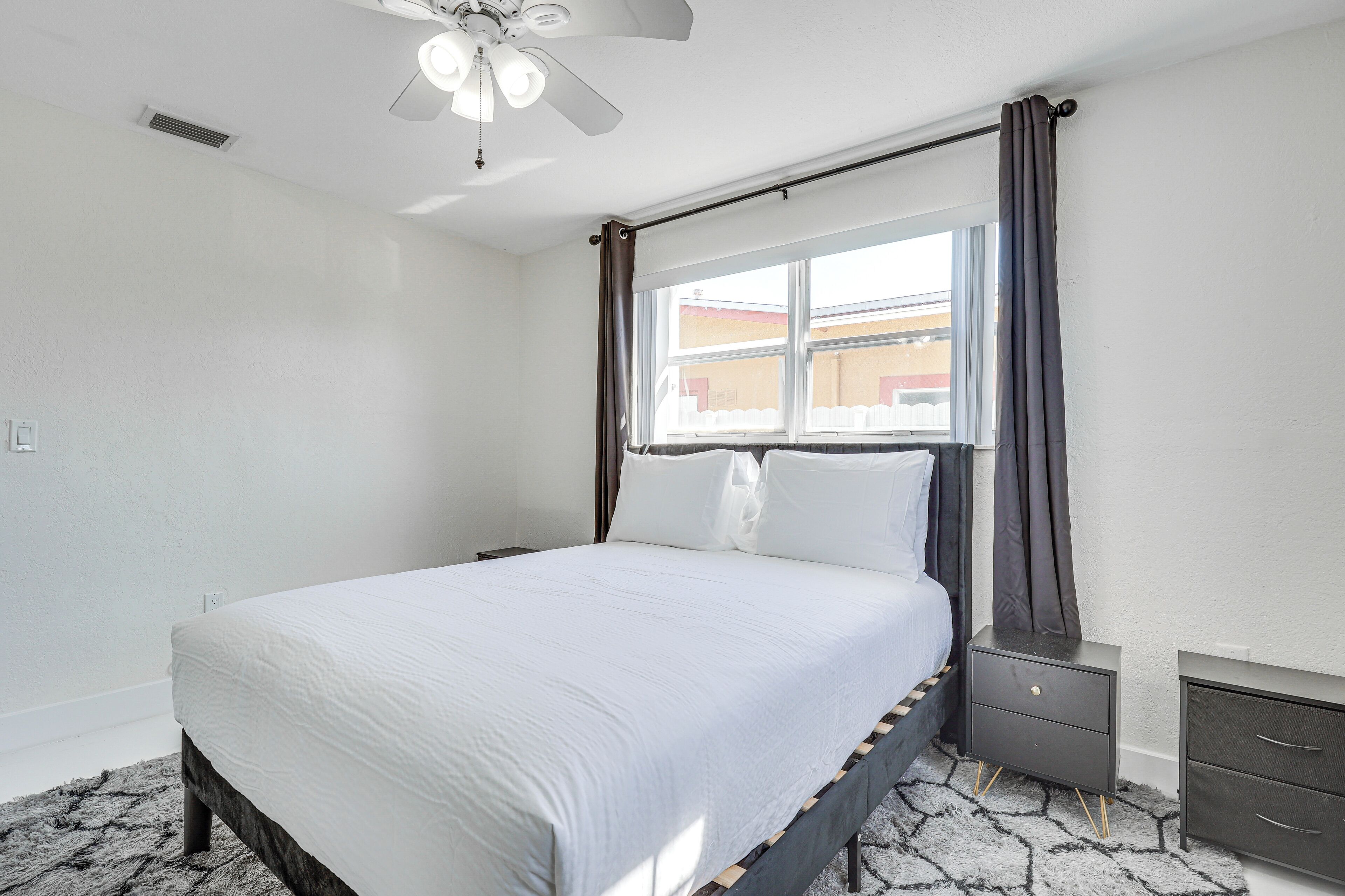 Une chambre à coucher comprenant un lit, une table de chevet, une commode, un ventilateur de plafond et une fenêtre avec des rideaux.