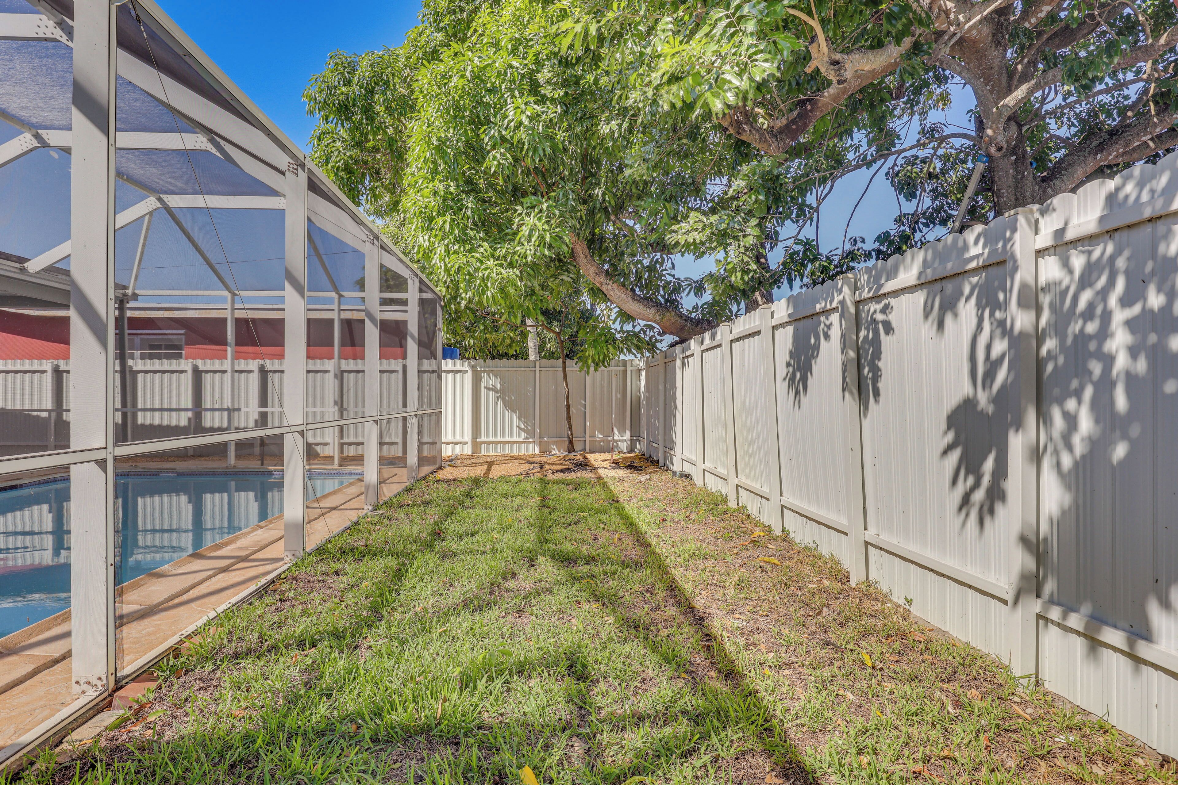 A backyard with a pool, a covered patio area, and a white fence.
