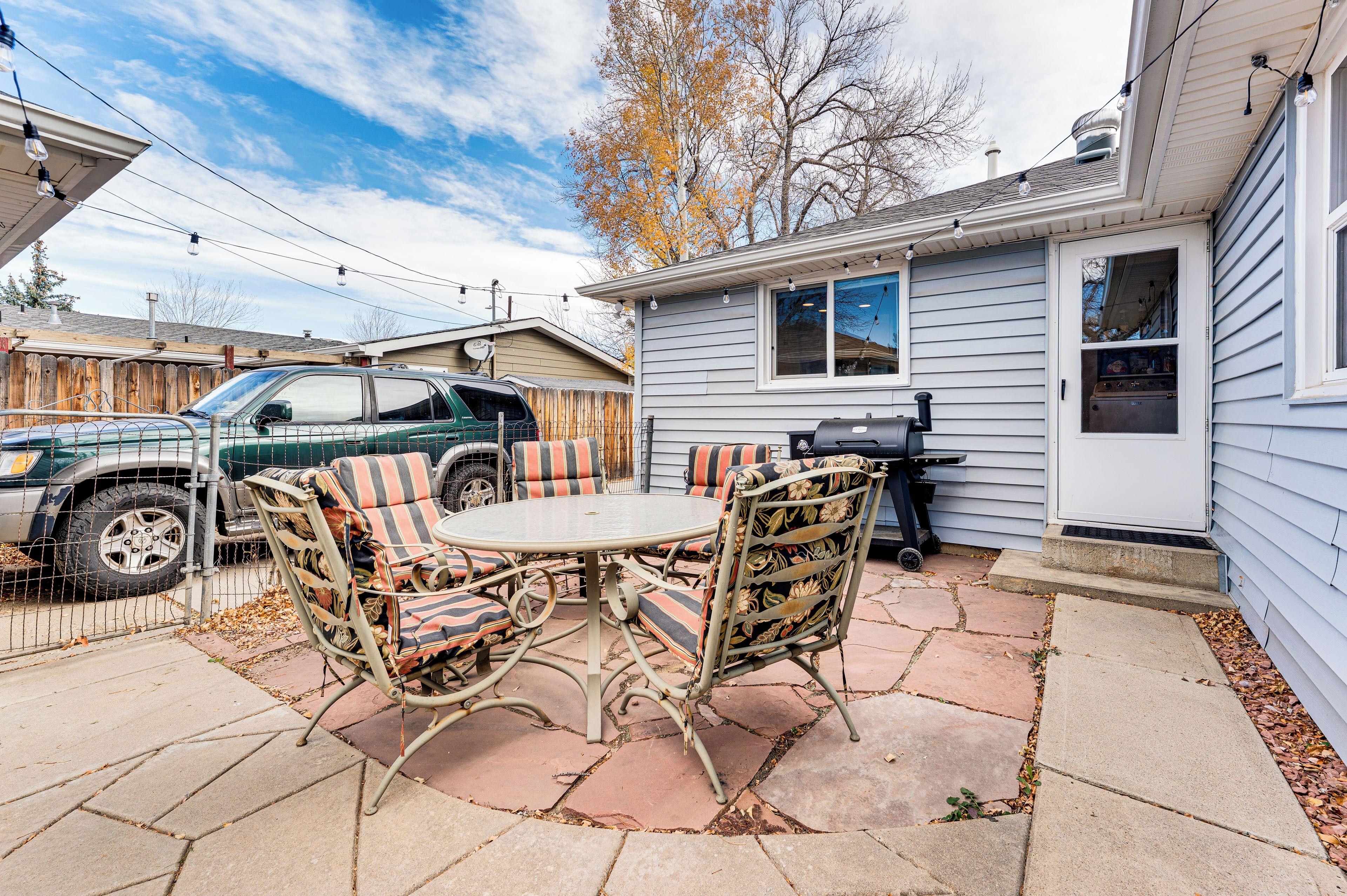A patio with a round table and chairs, a barbecue grill, and a vehicle parked in the background.