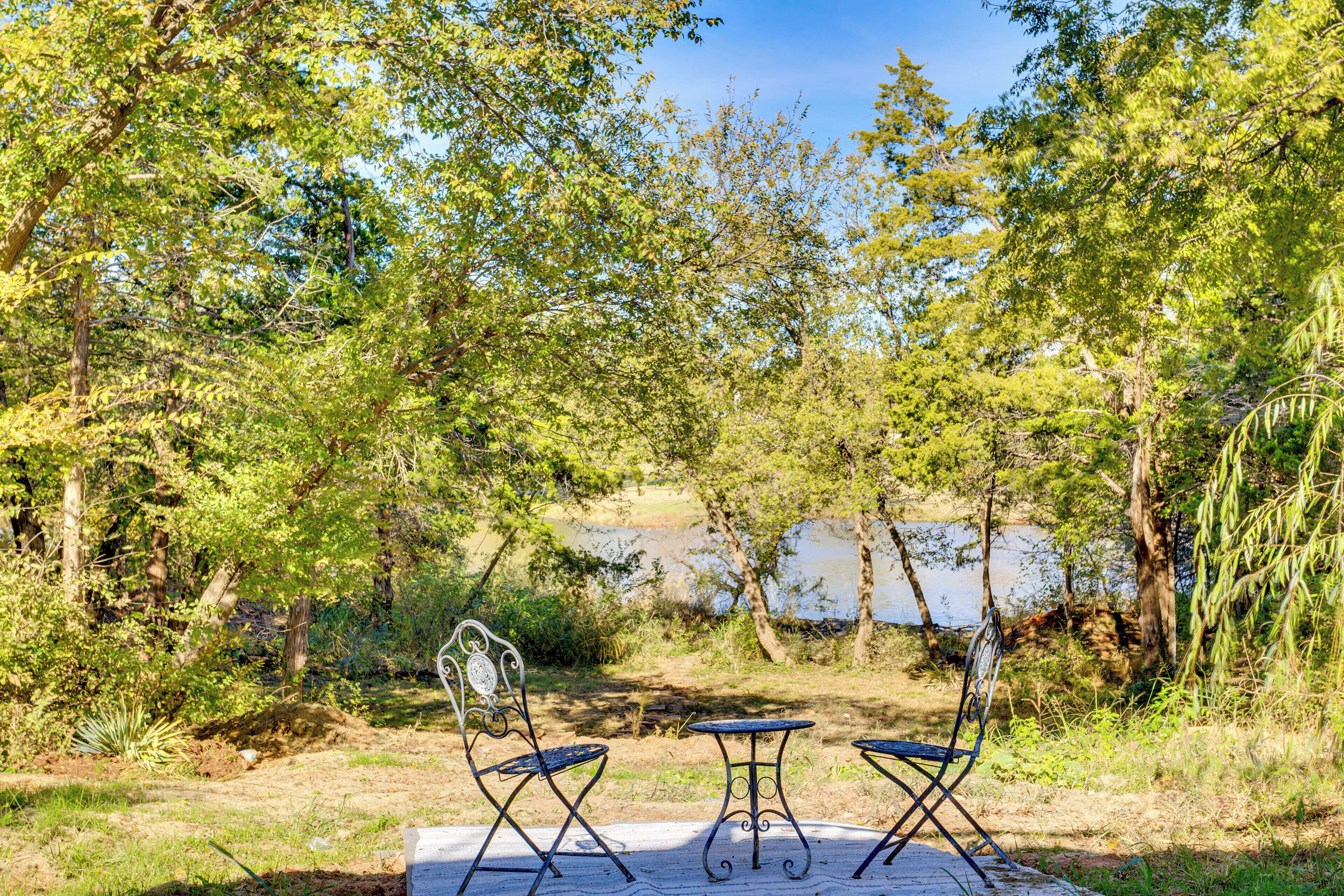 An outdoor seating area with metal chairs and a small table, surrounded by trees and a body of water.