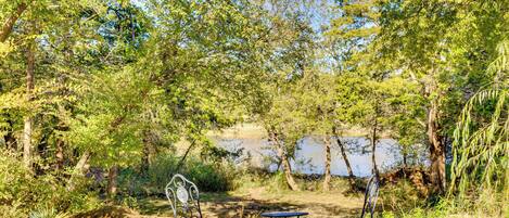 An outdoor seating area with metal chairs and a small table, surrounded by trees and a body of water.
