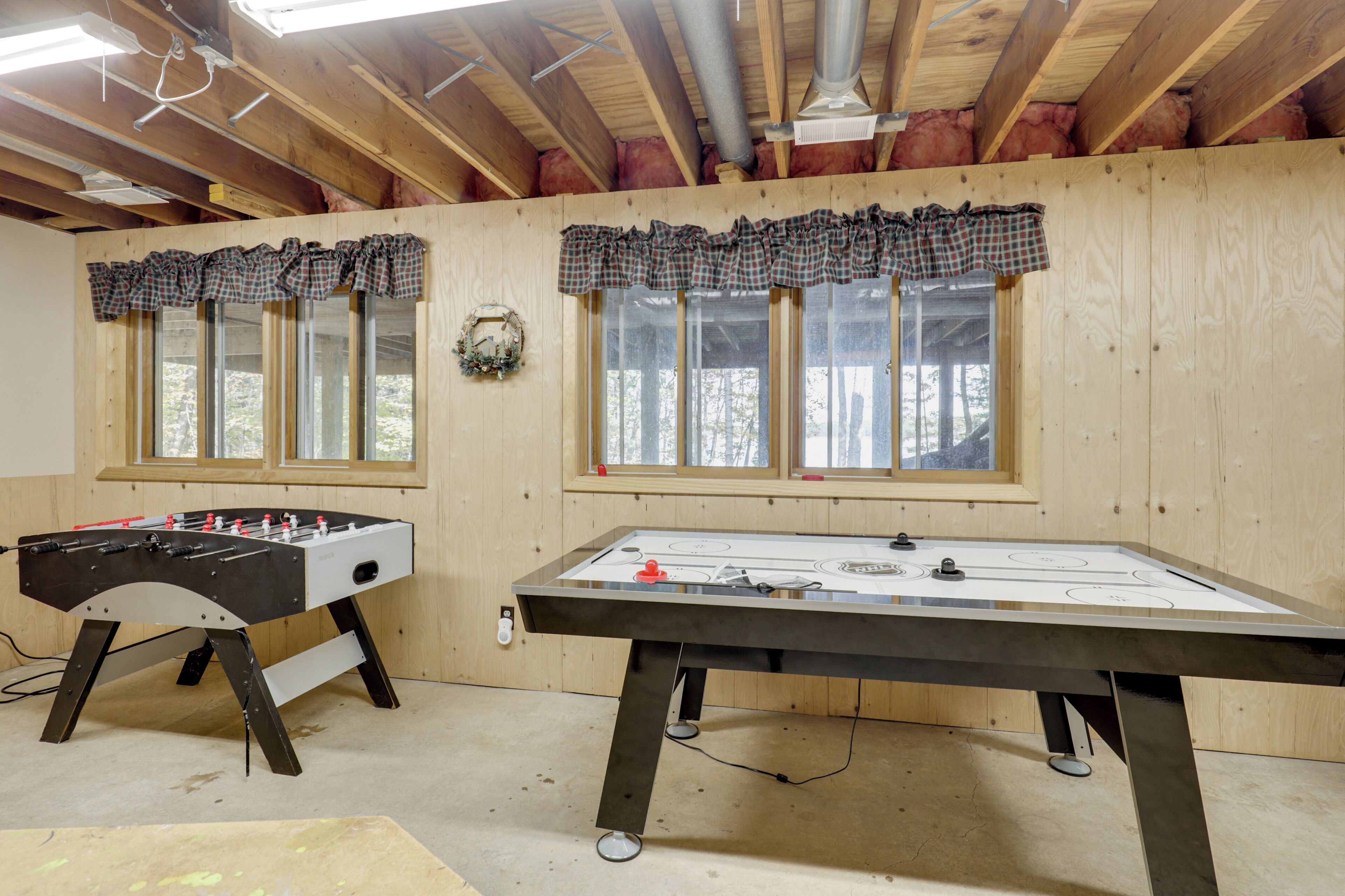 A room with two foosball tables, wooden walls, and windows with plaid curtains.