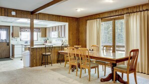 A dining area with a wooden table and chairs, a kitchen in the background, and large windows with curtains. - Land O' Lakes Cabin on Big Portage w/ Fire Pit (Land O' Lakes)
