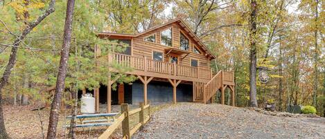 A wooden cabin with a balcony, surrounded by trees and a gravel driveway.