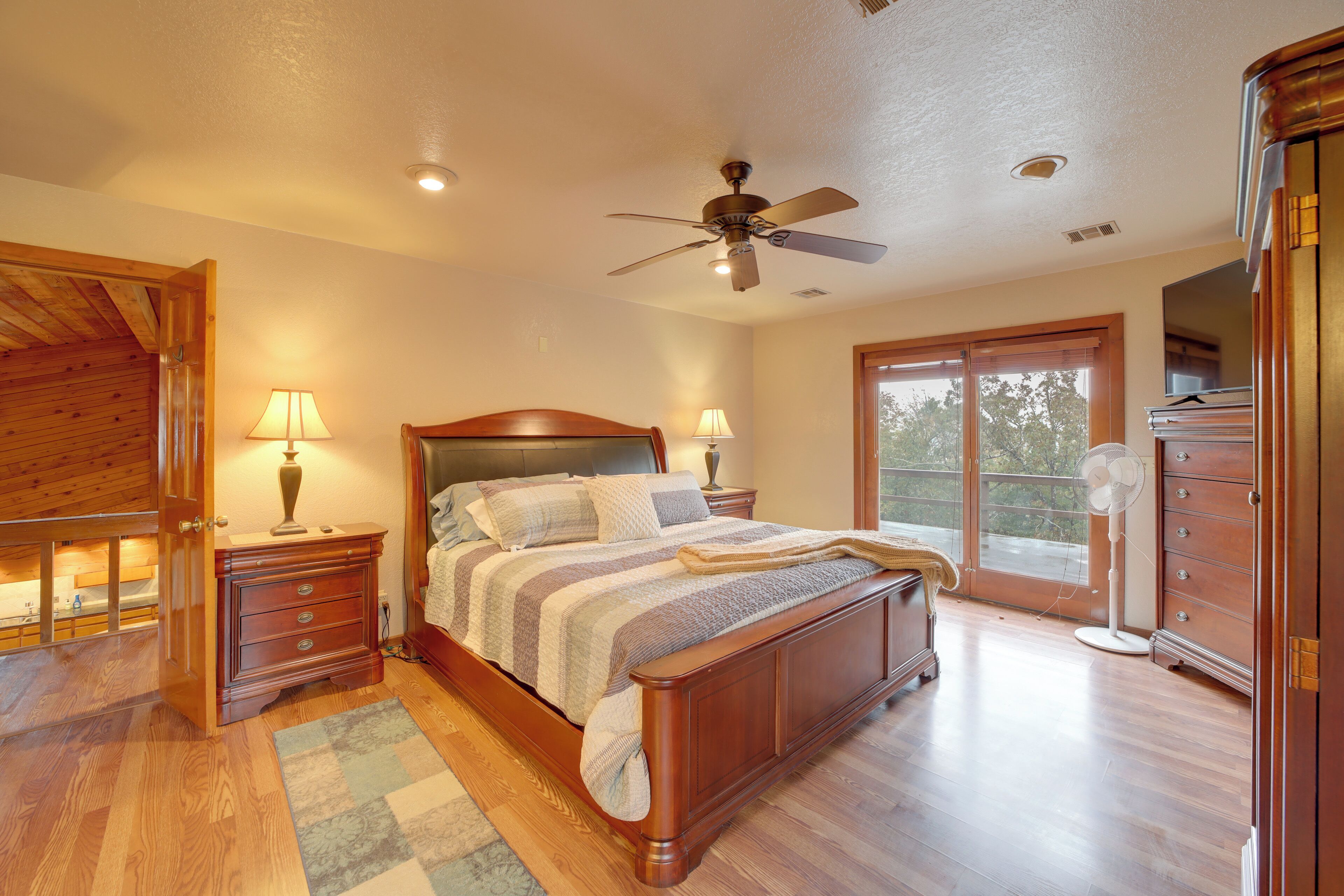 A bedroom with a wooden bed, two bedside tables with lamps, a ceiling fan, and a large window.