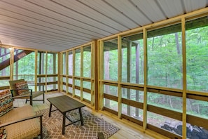 A covered outdoor seating area with wooden benches and a table, large windows, and a view of a forest. - Lake Access & Hot Tub! Serene Rockbridge Cabin (Amanda)