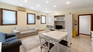 A living room with a sofa, a dining table with chairs, a kitchen area, and a clock on the wall.