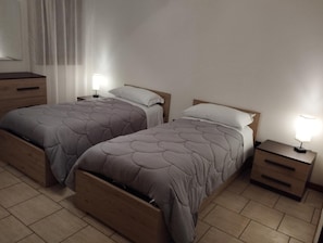 A bedroom with two beds, wooden headboards, bedside tables with lamps, and a window with curtains.