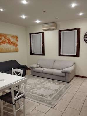 A living room with a gray sofa, a white table with chairs, a patterned rug, and two windows with curtains.