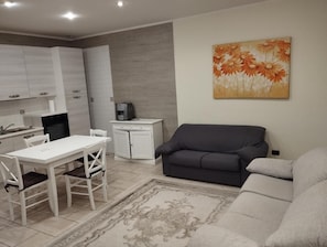 A modern living room with a grey sofa, a dining table with chairs, a kitchen area with cabinets, a coffee machine, and a decorative painting on the wall.