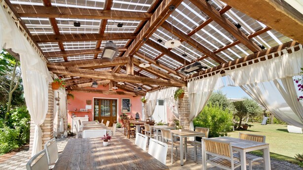An outdoor patio with wooden tables and chairs, a pergola with a wooden ceiling, and a view of the garden.