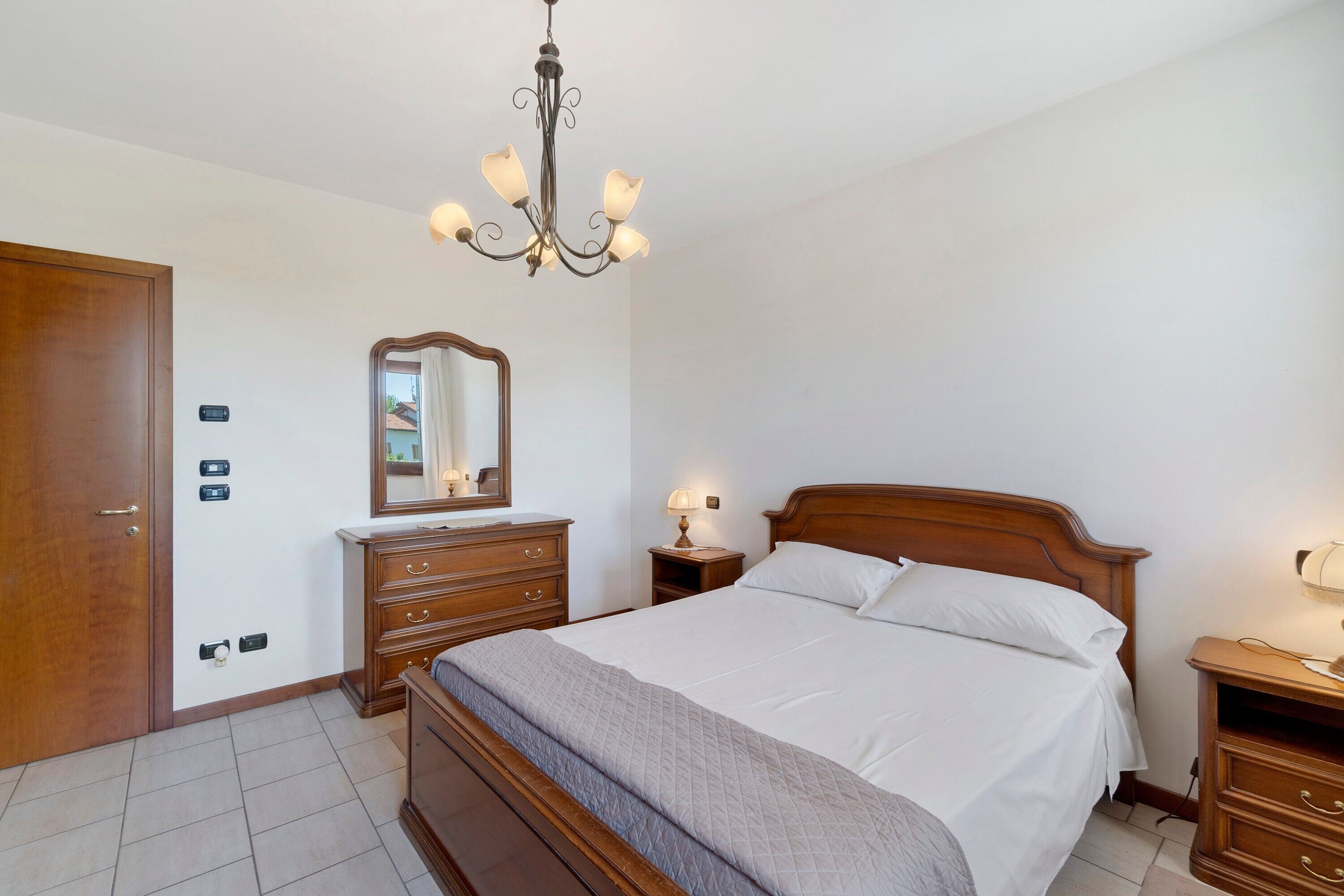 Un dormitorio con una cama de madera, una mesita de noche con lámpara, un aparador, un espejo y una araña.
