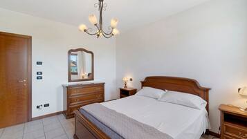 Un dormitorio con una cama de madera, una mesita de noche con lámpara, un aparador, un espejo y una araña.