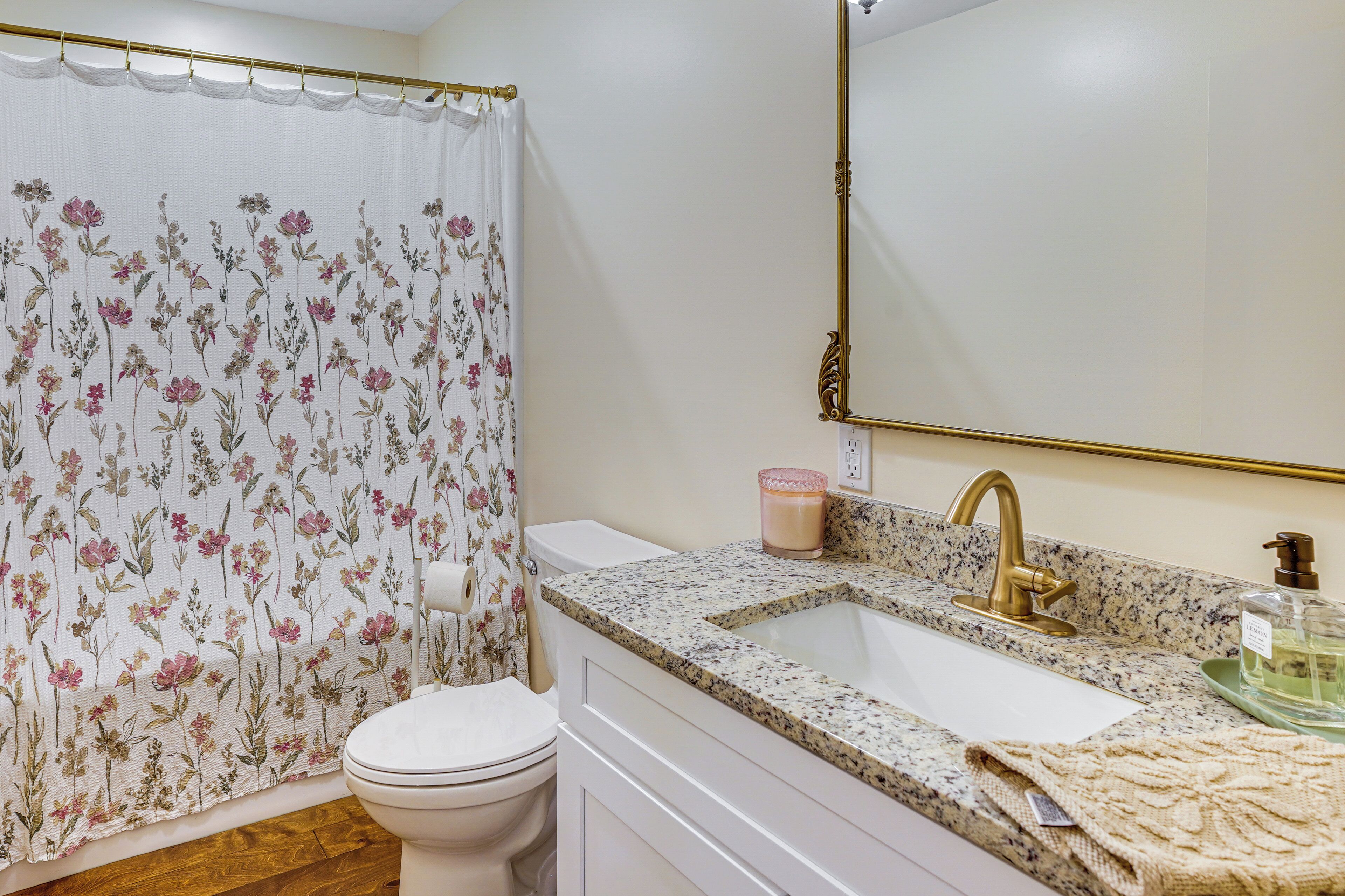 Ein Badezimmer mit floralem Duschvorhang, einer weißen Toilette, einem Waschbecken mit Granitplatte und goldenen Wasserhahn sowie einem Spiegel mit goldener Rahmung.