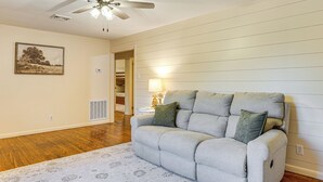 A living room with a gray sofa, a patterned rug, wooden flooring, a ceiling fan, a wall-mounted lamp, and a framed picture.