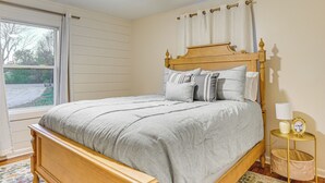 A bedroom with a wooden bed, a bedside table with a lamp, a window with curtains, and a view of the outdoors.