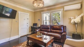 A living room with a brown leather couch, a wooden coffee table, a chandelier, and a large flat-screen TV mounted on the wall. - Easy Commute to Newark & Nyc! Extended Stay Unit (Irvington)