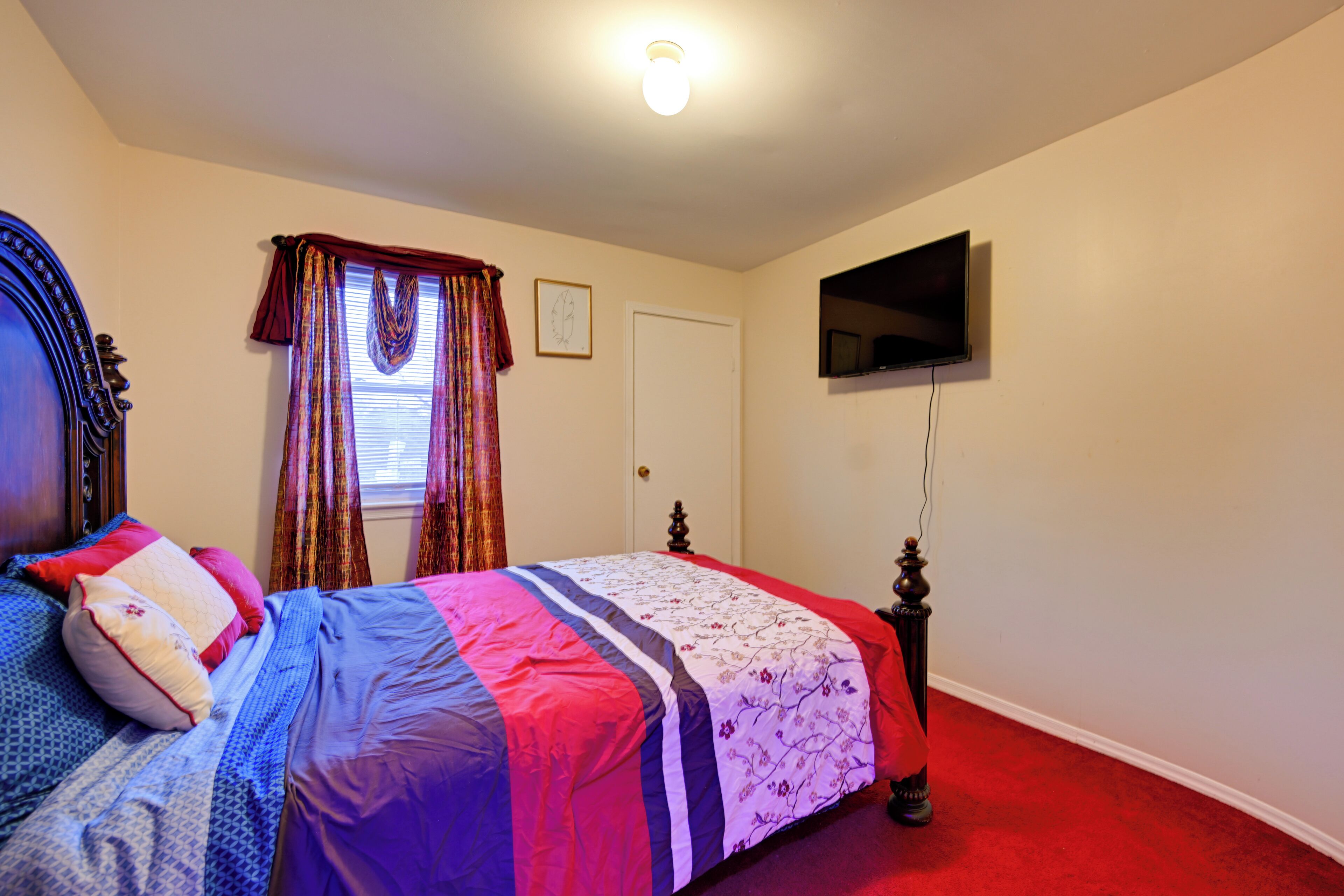 A bedroom with a bed, a television mounted on the wall, a door, and a window with curtains.
