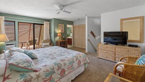 A bedroom with a bed, a television, a ceiling fan, and a wicker chair. - Maui Kamaole H-208 – Oceanview Poolside Condo Close to Beaches and Dining (Kihei)