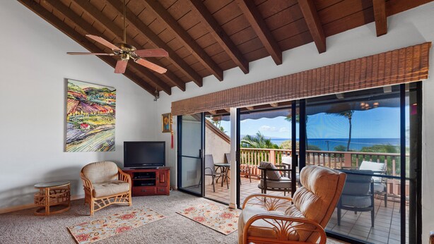 A room with a ceiling fan, a television, wicker furniture, a glass door leading to an outdoor area, and a painting on the wall. - Maui Kamaole H-208 – Oceanview Poolside Condo Close to Beaches and Dining (Kihei)