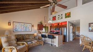 A spacious living area with a high ceiling, a central island with bar stools, a sofa, and a dining table. - Maui Kamaole H-208 – Oceanview Poolside Condo Close to Beaches and Dining (Kihei)