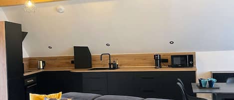 En moderne stue med en hjørnesofa, et køkkenområde med mikroovn og kaffemaskine, samt synlige træbjælker i loftet.