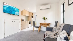 A compact living space with a kitchenette, a dining area with a table and chairs, a grey sofa with a blanket, and a flat-screen TV mounted on the wall. - Cozy Central Loft | Walk to CBD & Stadium (Christchurch)