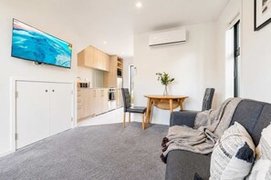 A compact living space with a kitchenette, a dining area with a table and chairs, a grey sofa with a blanket, and a flat-screen TV mounted on the wall. - Cozy Central Loft | Walk to CBD & Stadium (Christchurch)
