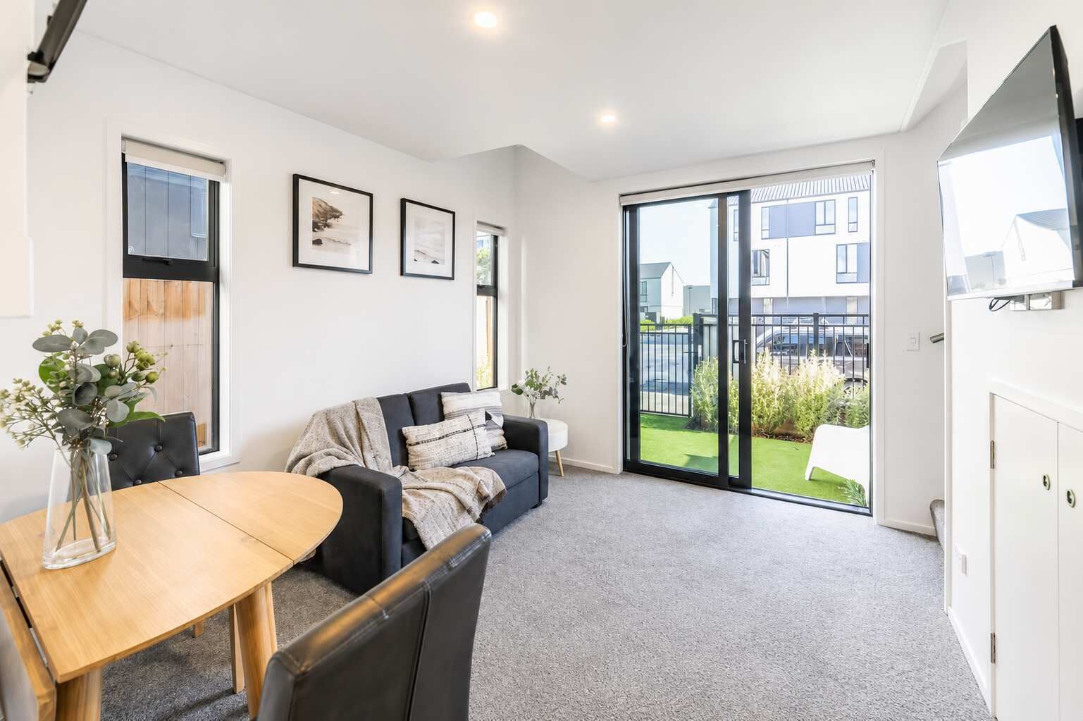Una sala de estar moderna con un sofá, una mesa de comedor y vistas a una zona residencial a través de amplios ventanales corredizos.