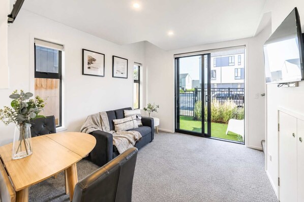 A modern living room with a sofa, a dining table, and a view of a residential area through large sliding glass doors. - Cozy Central Loft | Walk to CBD & Stadium (Christchurch)