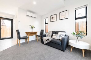 Ein modernes Wohnzimmer mit einer Couch, einem Esstisch und Stühlen. An der Wand befinden sich gerahmte Bilder und ein Fenster mit Jalousien.