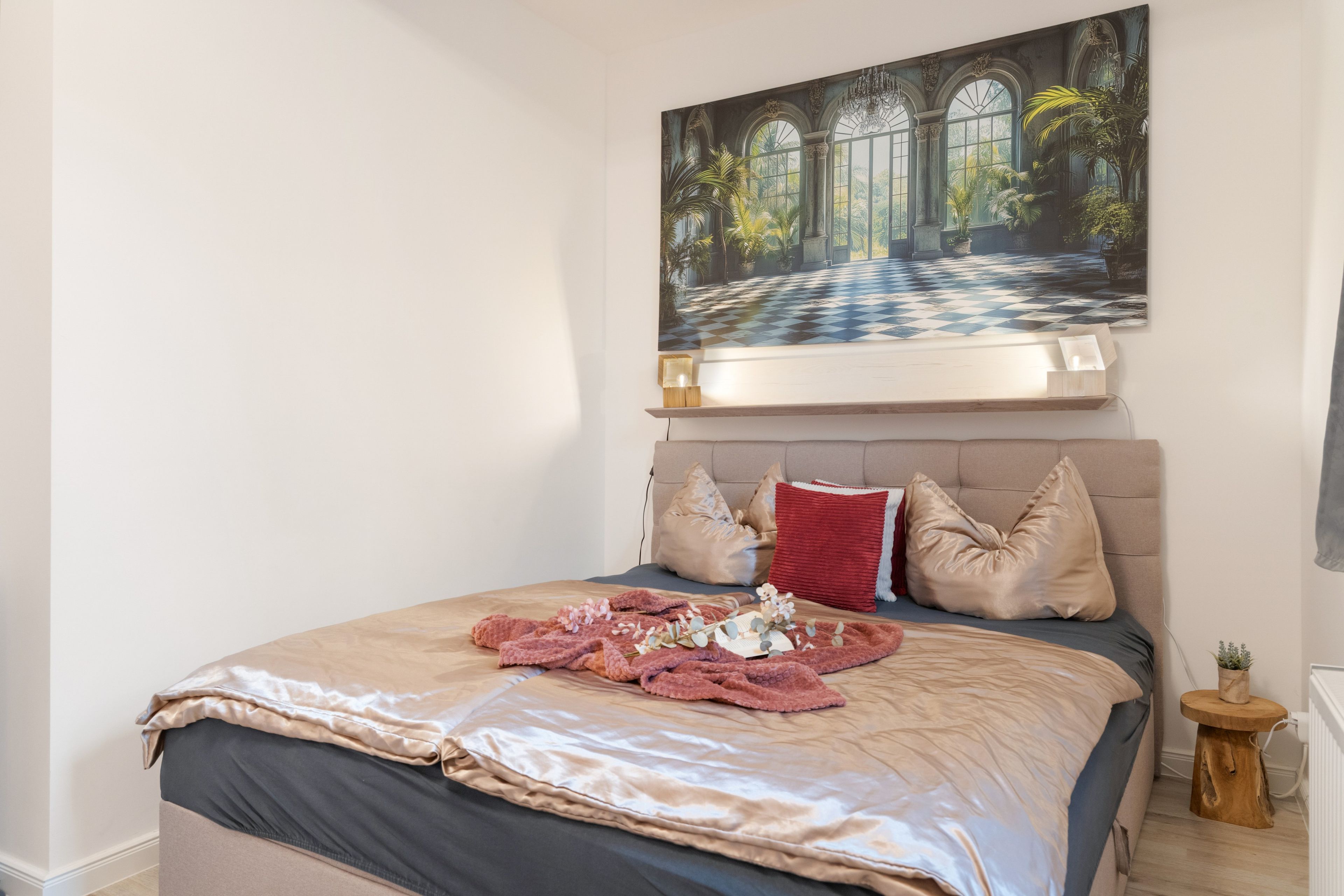 A bedroom with a large bed, a painting above it, and a small wooden side table.