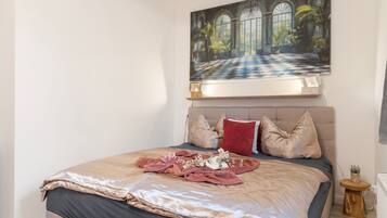 A bedroom with a large bed, a painting above it, and a small wooden side table.