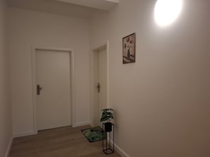Un passadís amb una porta blanca, una planta petita, un quadre amb marc i un lluminari a la paret.