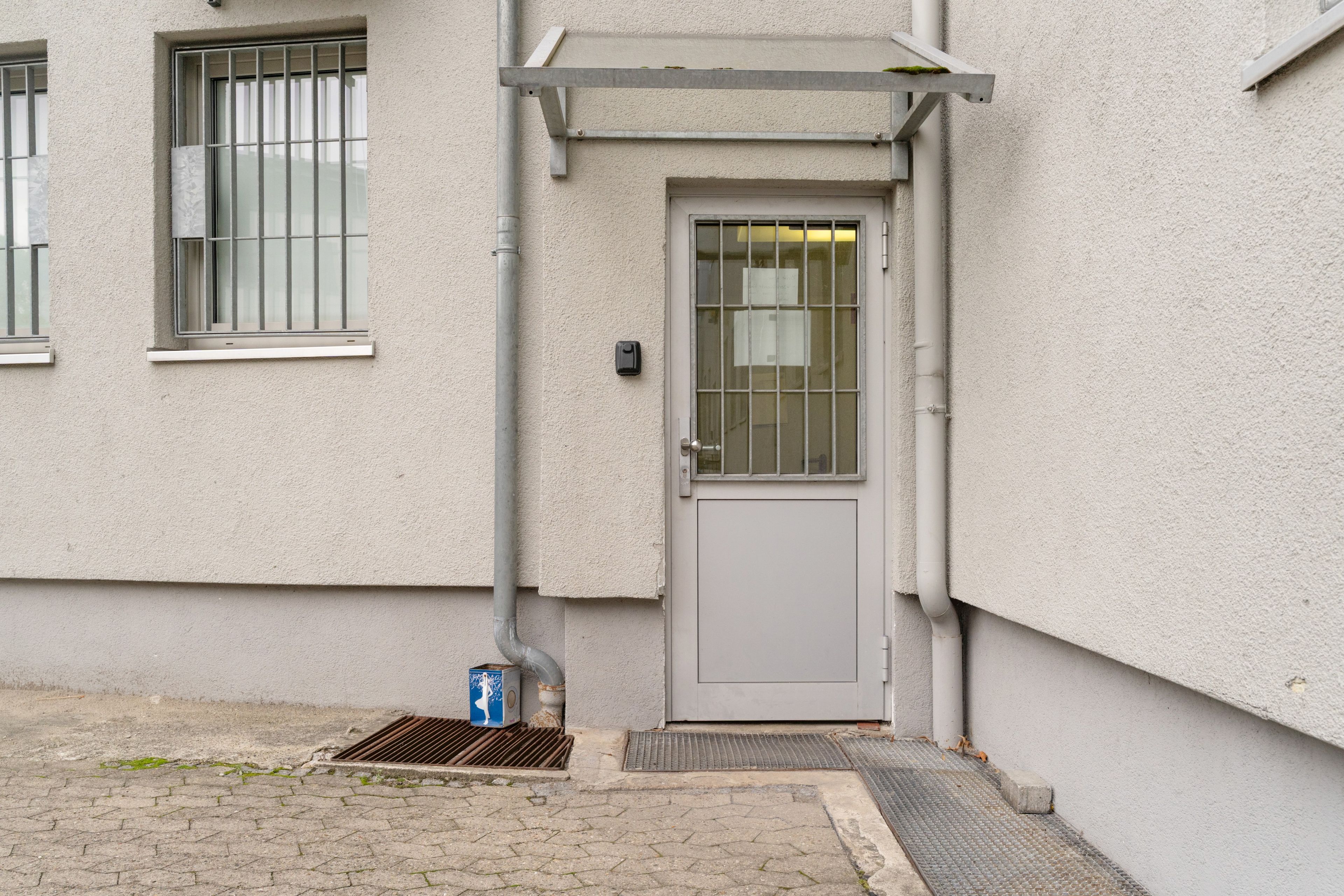 Une entrée d’immeuble avec une porte grise, une petite marche et une grille métallique.