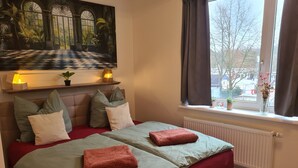 Een slaapkamer met een geruit vloerkleed, een bed met rode beddengoed, een raam met gordijnen en een schilderij boven het bed.