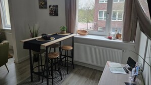 Een kleine keuken met een eethoek, een radiator, een raam met gordijnen en uitzicht op een bakstenen gebouw.