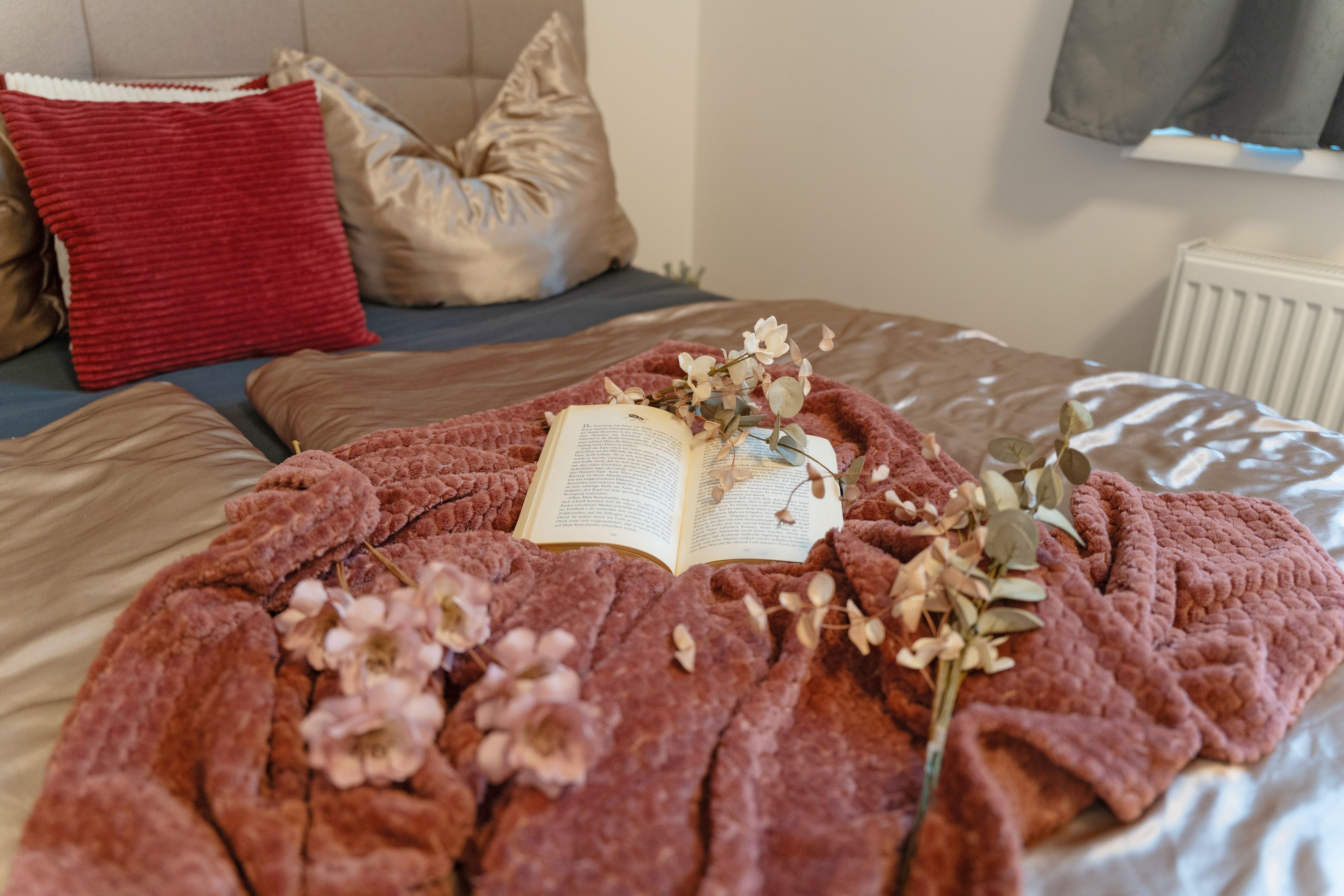 A bed with a red blanket, pillows, and an open book with flowers on it.