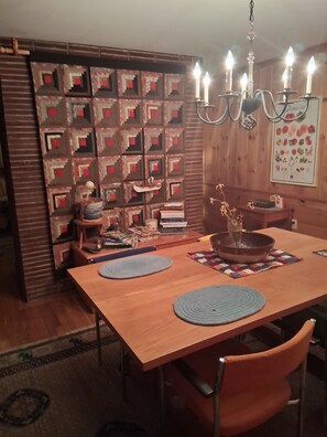 A dining area with a wooden table, two blue placemats, a chandelier, and a wall decorated with framed pictures. - Tranquil 3-bedroom bungalow with AC in Moultonborough. On the lake. (Moultonborough)