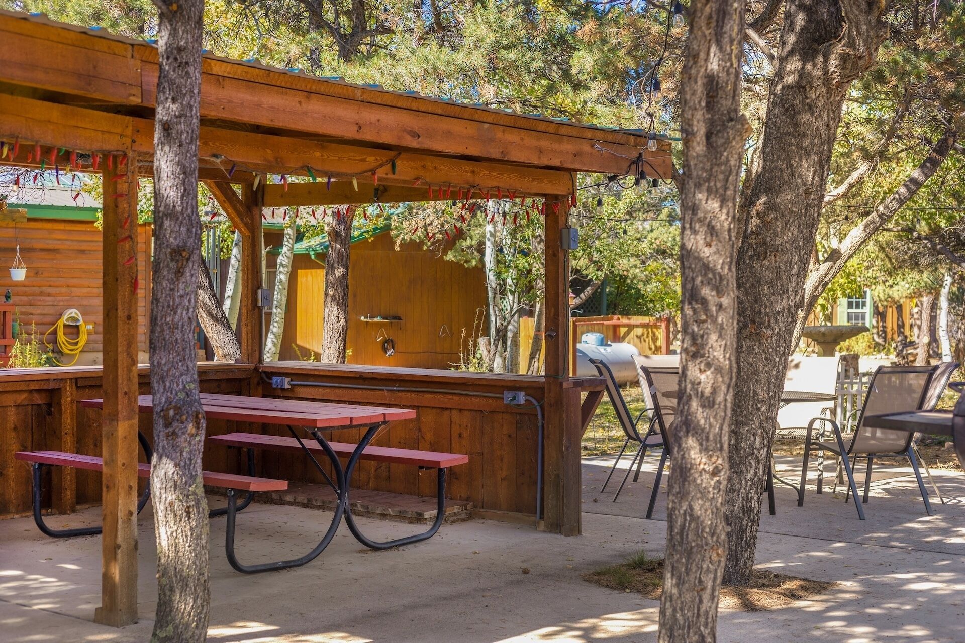 A wooden pergola with picnic tables and chairs, surrounded by trees.