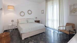 Un dormitori amb llit, taules de nit, una làmpada, una cadira, una cistella de vimet i una finestra amb cortines.
