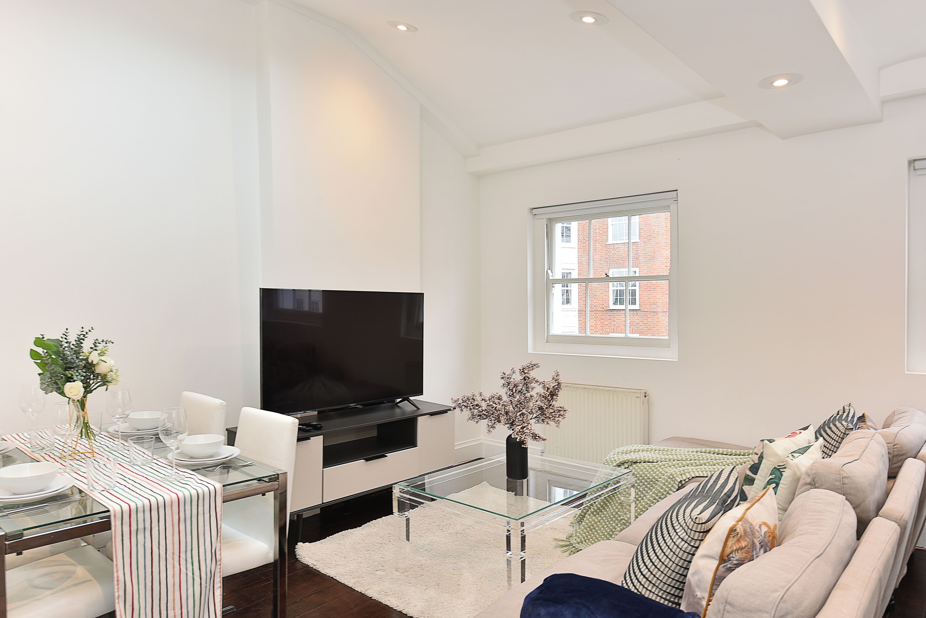 A modern living room with a glass dining table, a flat-screen TV, a sofa, and a large window with a view of a brick building.